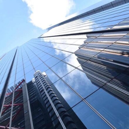 Reflections of high rise buildings appearing in the glass windows of another high rise building with a partly cloudy sky above.