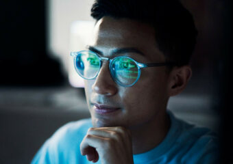 Asian man with glasses works on coding, with reflection of monitor appearing on his glasses.