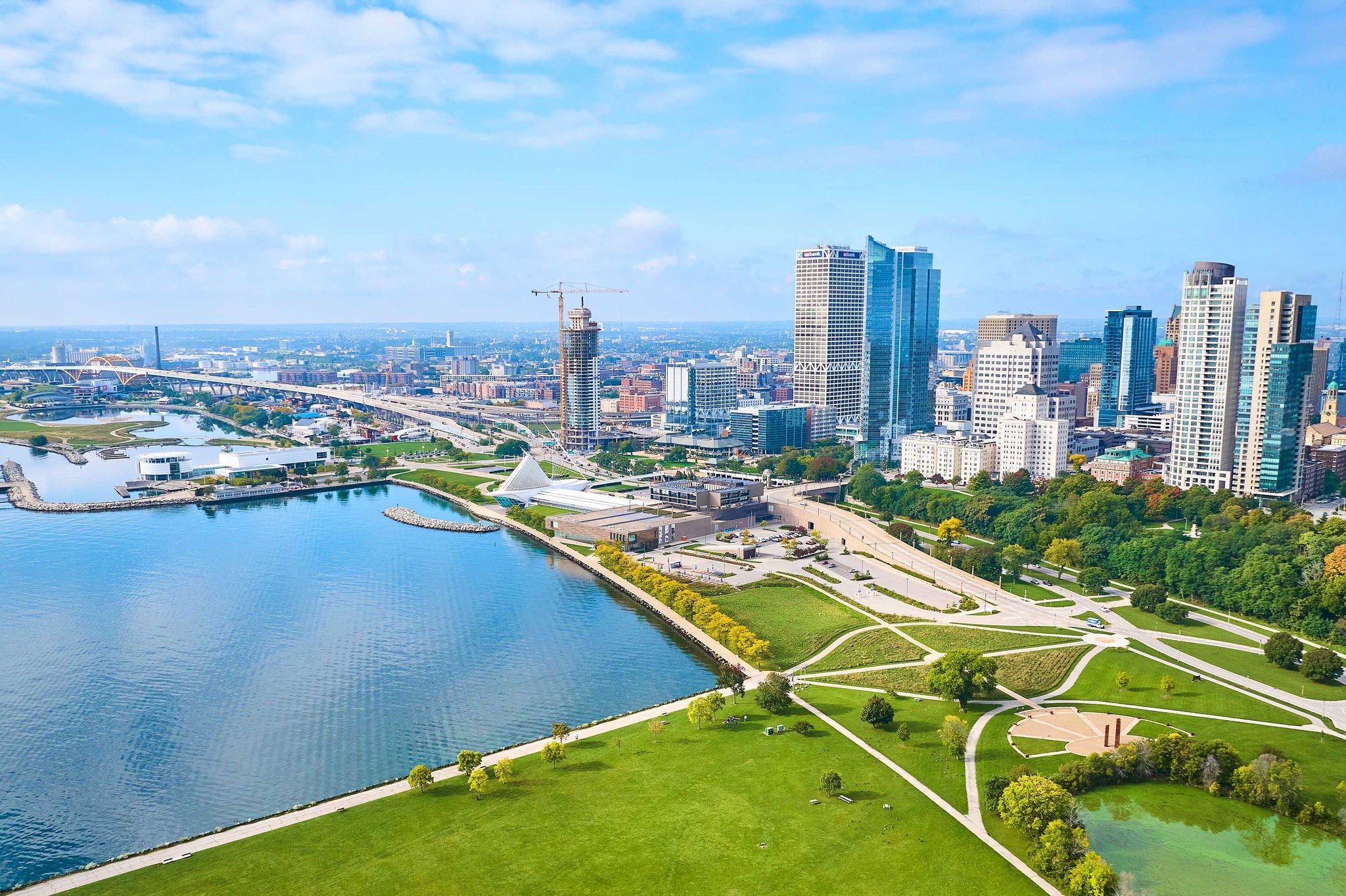 Drone view of Milwaukee's vibrant cityscape, showcasing the contrast between lush parklands and modern high-rise buildings, with iconic Quadracci Pavilion and Lake Michigan in view.