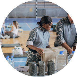 A woman stands at a work bench assembling a box of water bottles and other emergency supplies