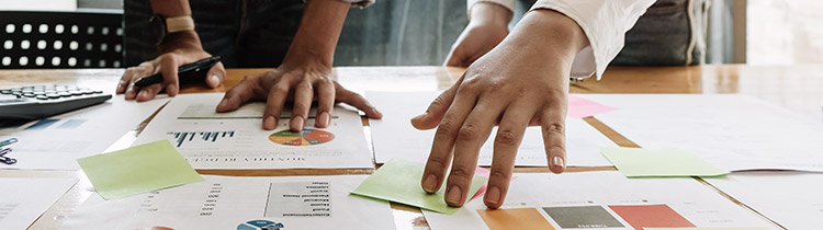 Close-up of a table top with papers and graphs and sticky notes, with two people standing behind it and collaborating