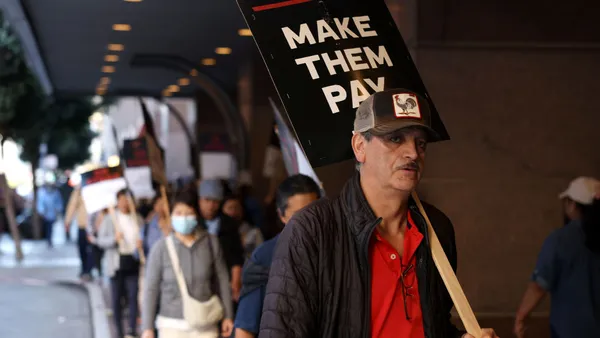 A worker on a picket line holds a sign reading "make them pay."