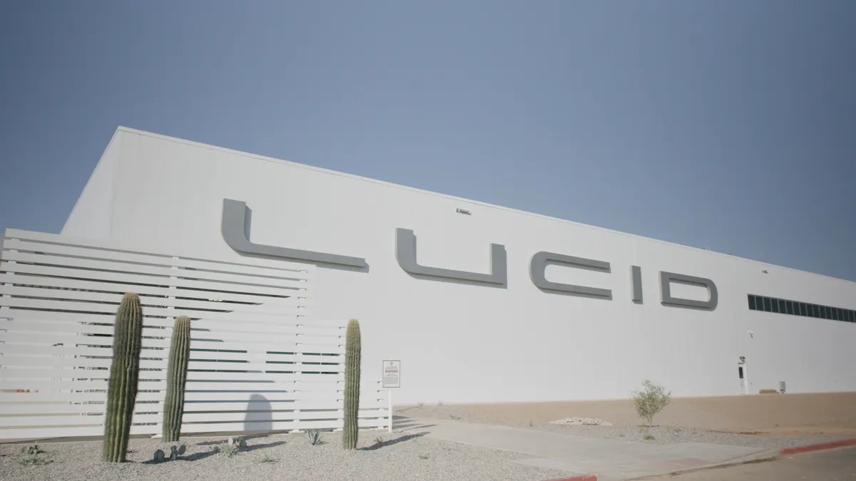 White building with Lucid in big gray letters and dark windows next to it on a sunny day in the desert. Three green cacti are planted in front of the building.