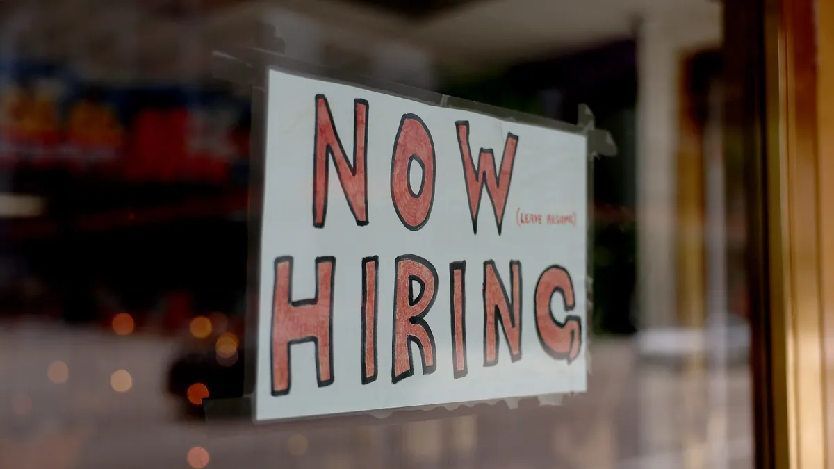 A handwritten "now hiring" sign hangs in a shop window.