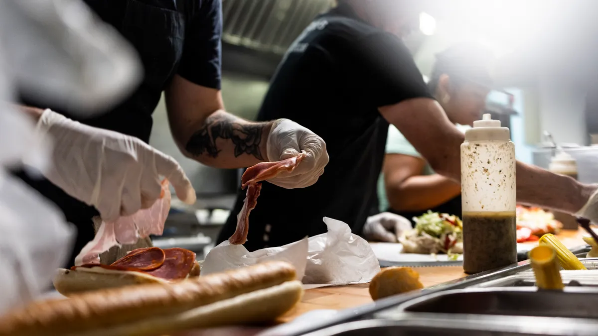 Sandwiches sit on a counter as workers as ingredients.