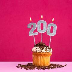 A birthday cupcake with lit silver candles saying '200'. The cupcake has white icing and chocolate sprinkles. The background of the image is pink. Image by Luis Echeverri Urrea via Shutterstock.