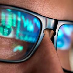 A close-up of the face of a man wearing dark-rimmed glasses. The glasses show the reflection of the screen he is looking at. Image by Ground Picture via Shutterstock.