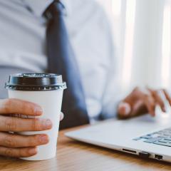 A man in a suit and tie looking at something on a laptop. He is holding a paper cup in his right hand. Image by Lisa Fotios via Pexels.