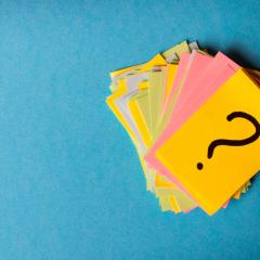 A pile of pieces of yellow, green, blue and pink paper against a blue background. The top piece of paper is bright yellow and has a question mark on it. Image by SNeG17 via Shutterstock.