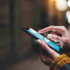 Close up of the hands of a woman using a smartphone. She has brown skin and is wearing a chunky knit ochre jumper. The background is blurred and quite dark. Image by A_B_C via Shutterstock.