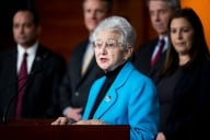 Virginia Foxx, in a bright blue blazer, stands behind a podium.