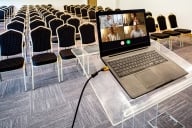 A computer with four people on the screen sits on a podium before a room of empty chairs 