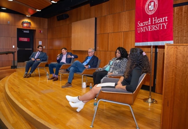 On a large wooden stage, four trustees and a student sit in wooden chairs facing an audience for a Q&A-style discussion.