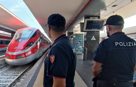 Poliziotti ritratti a mezzobusto, uno di spalle l'altro di profilo, sul bordo di un binario impegnato da un treno