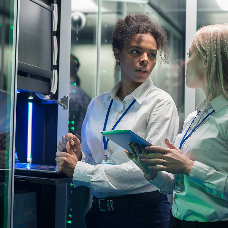 Medium shot of two women working in a data center with rows of server racks and checking the equipment and discussing their work