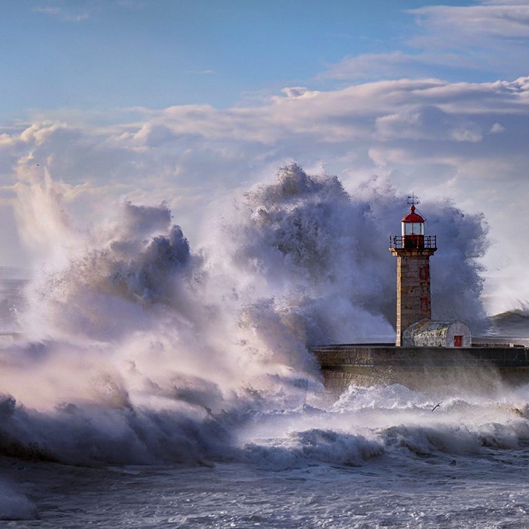 Lighthouse at sea with choppy waters and waves hitting the lghthouse.