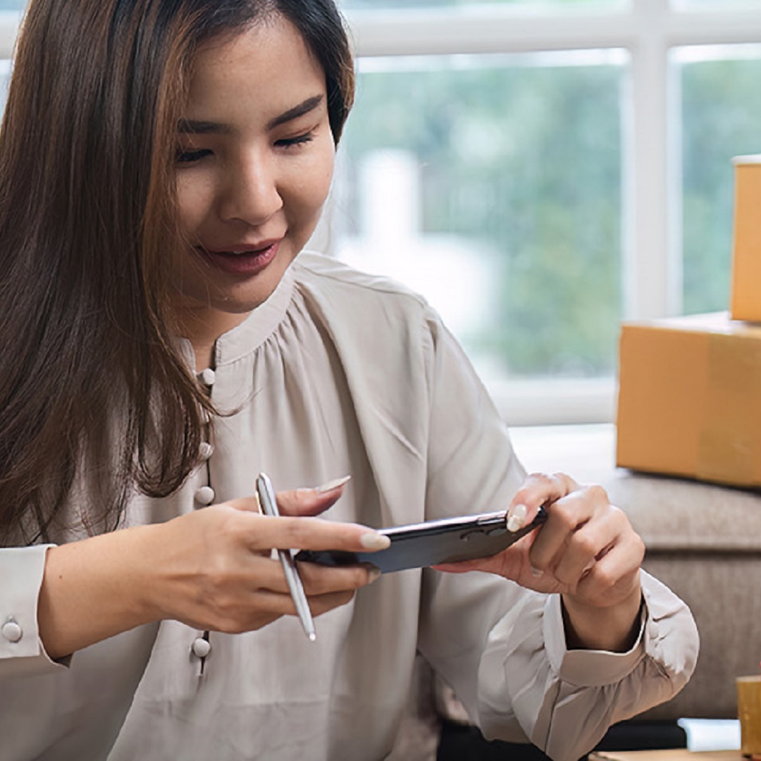 A young woman managing her small online sales business from home, surrounded by packages, using her smartphone to take product photos.