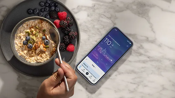 A person eats oatmeal with berries, and a phone on the counter shows their current glucose levels.