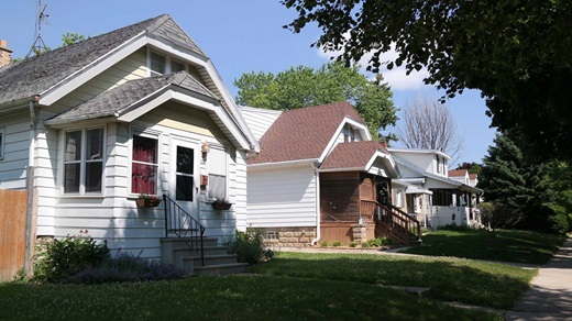 On a sunny summer day, several small, older, bungalow-style single-family homes appear along a tree-lined street in an urban neighborhood. The houses are tidy, but there are signs of deferred maintenance that suggest the homeowners might have limited means to make repairs. The house in the foreground has old, curling roof shingles, for example.