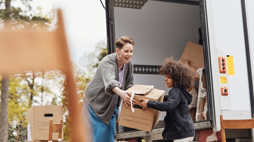 On a cloudy day, a woman and her young son unload moving boxes from the back of a white moving van. Dining room chairs and other moving boxes appear in the foreground and background. The woman is wearing a gray cardigan and denim skirt and her son is wearing a dark gray hoodie.