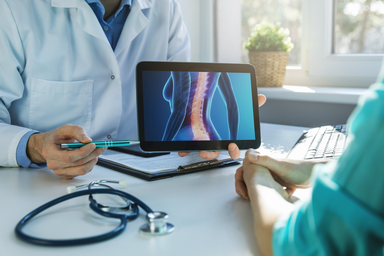 A patient in a green top sits at a desk across from a doctor in a white lab coat. The doctor is showing the patient a 3D drawing of a human spine on a tablet. Their faces are out of frame.