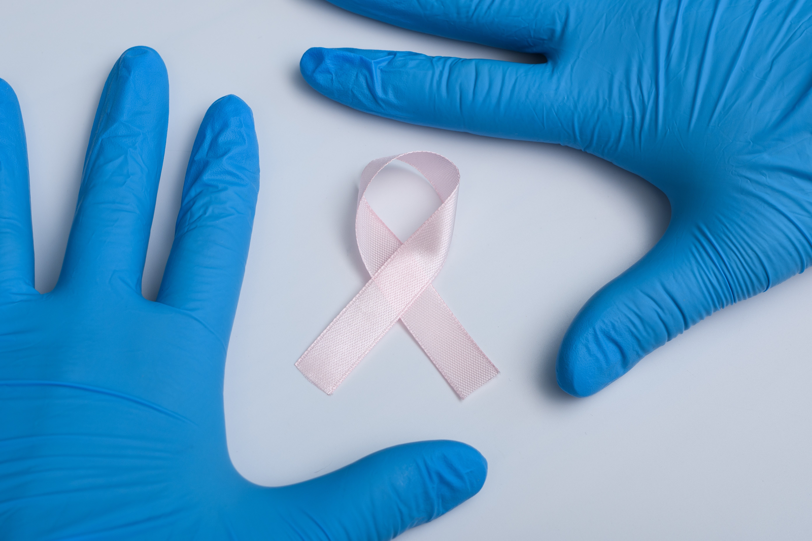 Two hands wearing blue surgical gloves frame a pink breast cancer ribbon on a flat white surface.