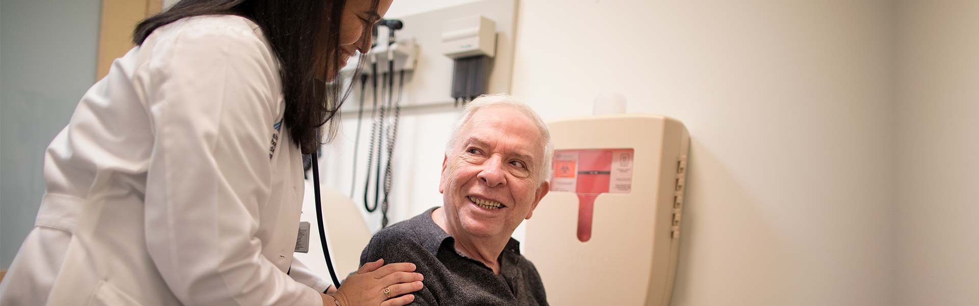 Female doctor using a stethoscope to evaluate a seated male geriatric patient 