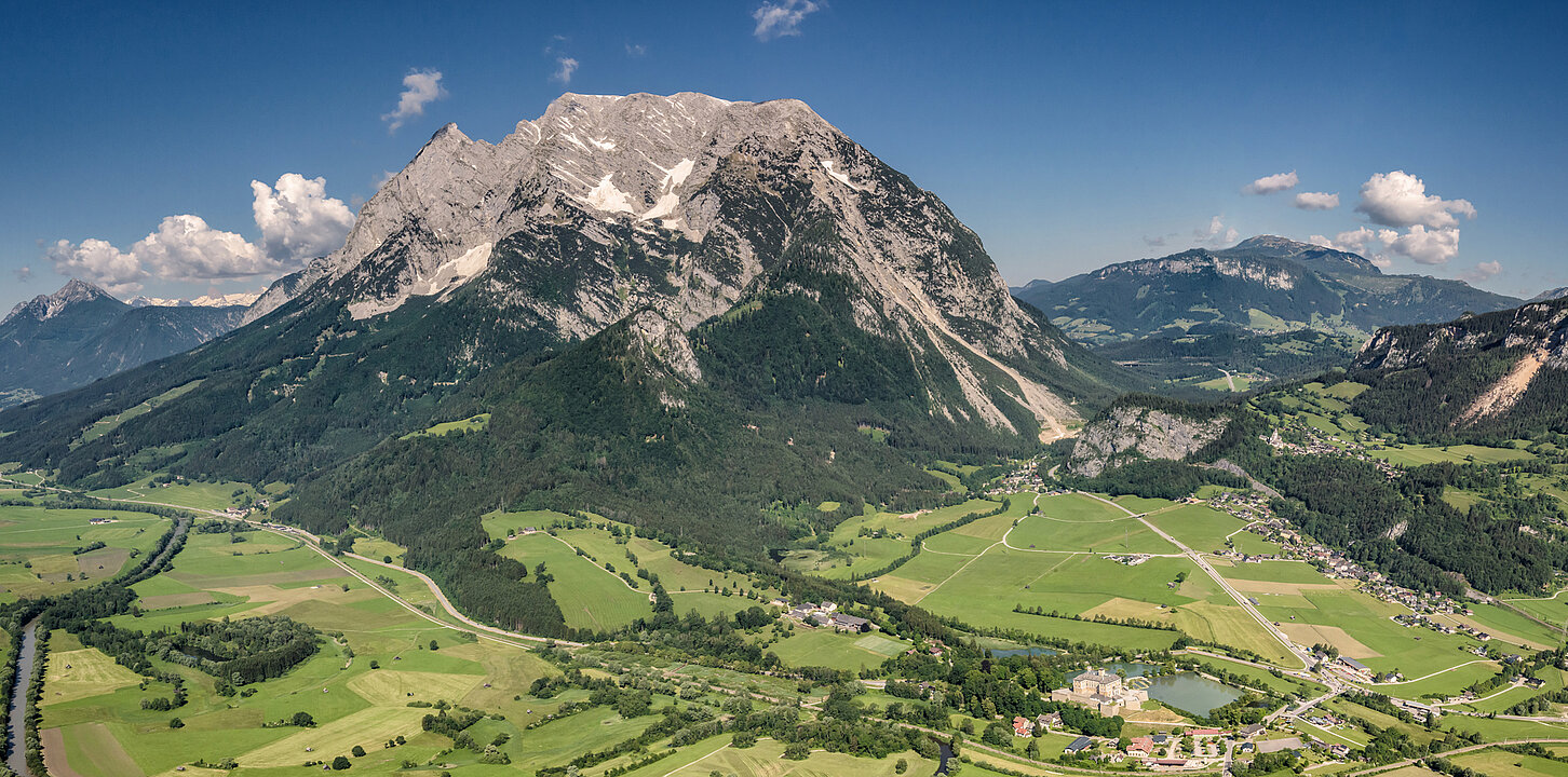 Aerial view of Grimming mountain with blue sky and green meadows. In the lower right corner of the picture you can see the castle.