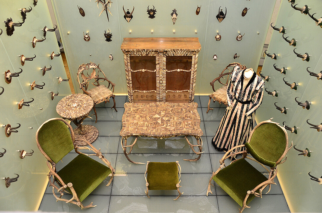 In a showcase are several pieces of furniture and a mannequin wearing a striped dress. The furniture is covered with green velvet and is otherwise made entirely of antler and horn.