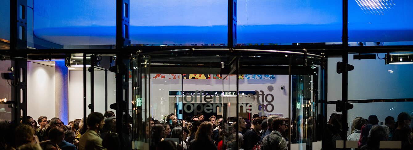 The Kunsthaus Graz is photographed from the outside at night. The blue-lit foyer of the Kunsthaus can be seen through the glass façade. A large crowd gathers in the foyer and a choir stands above the crowd on a balcony-like bridge.