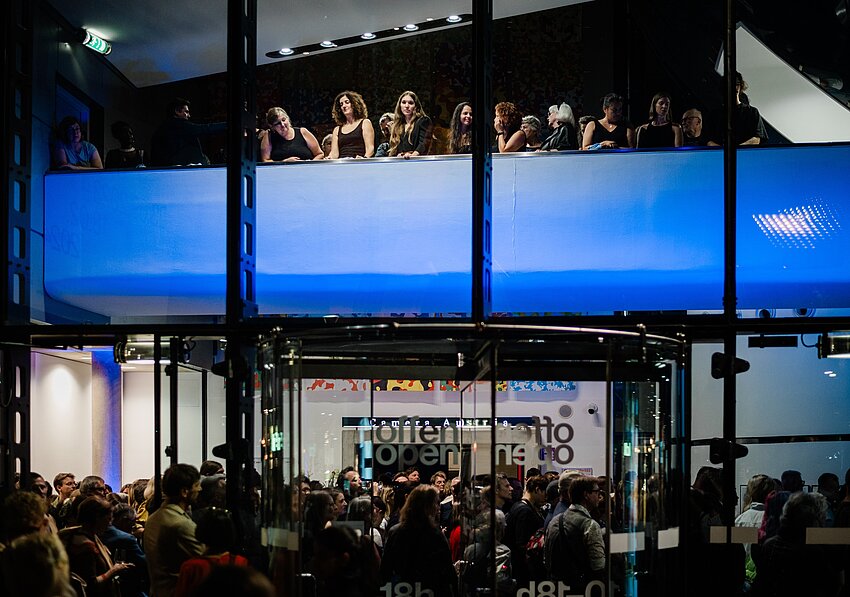 The Kunsthaus Graz is photographed from the outside at night. The blue-lit foyer of the Kunsthaus can be seen through the glass façade. A large crowd gathers in the foyer and a choir stands above the crowd on a balcony-like bridge.