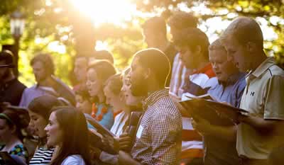 A group of students singing while sunshine pours through the trees above them