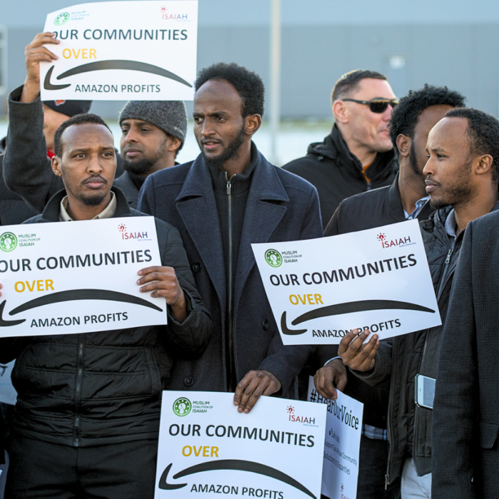 A group of men of color participate in an action. Their signs read, "Our Communities Over Amazon Profits." - Good Jobs Economy National Employment Law Project