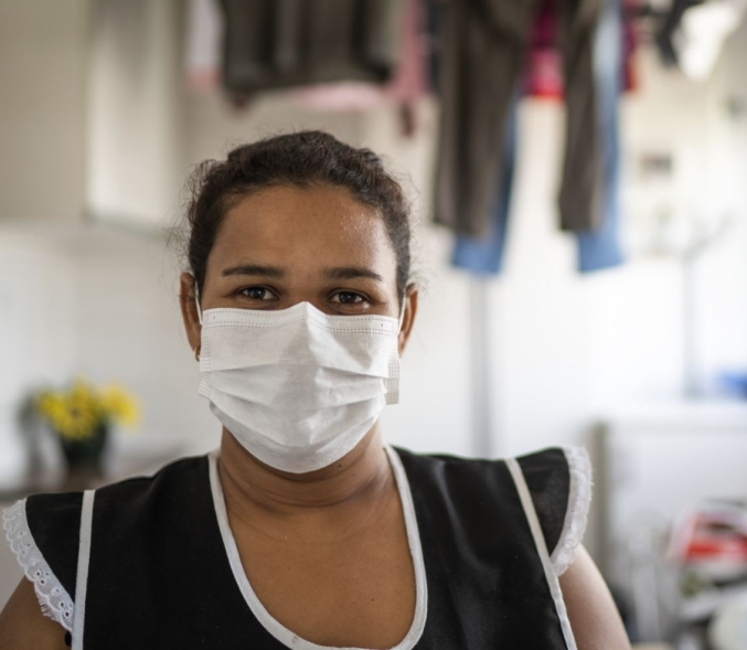 A Latinx woman housekeeper wears a white paper mask and black apron in a home with hanging laundry