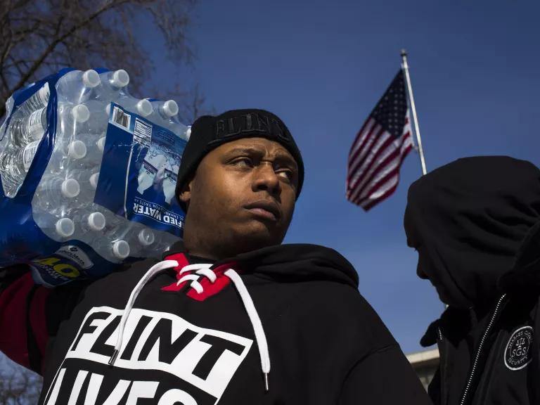 A man carries a large case of bottled water on his right shoulder