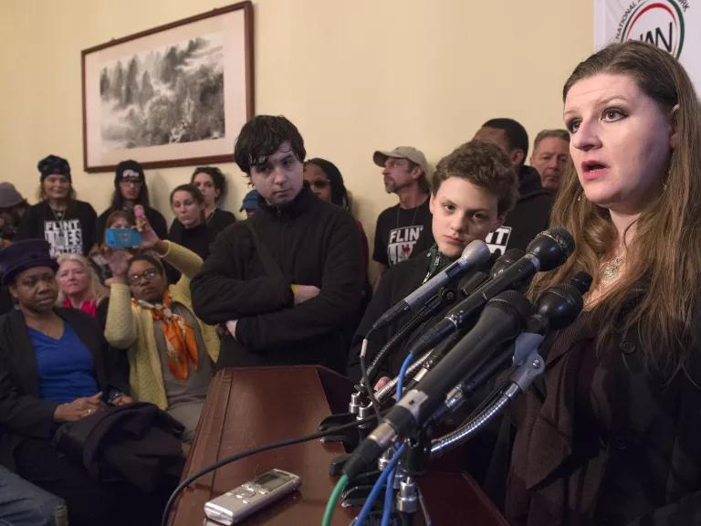 A woman speaks at a lectern into a cluster of microphones, with a crowd of people looking on.