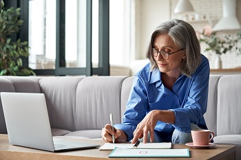 woman with glasses working at home with a laptop and notebook.