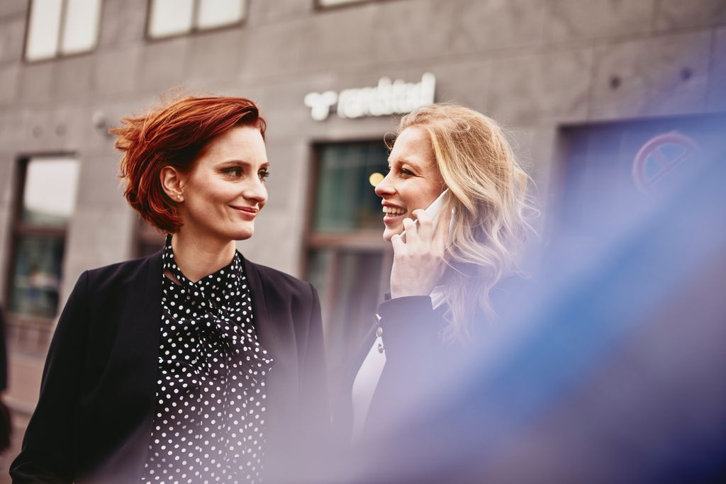 Two smiling woman, talking and walking around the city. One woman is on the phone.