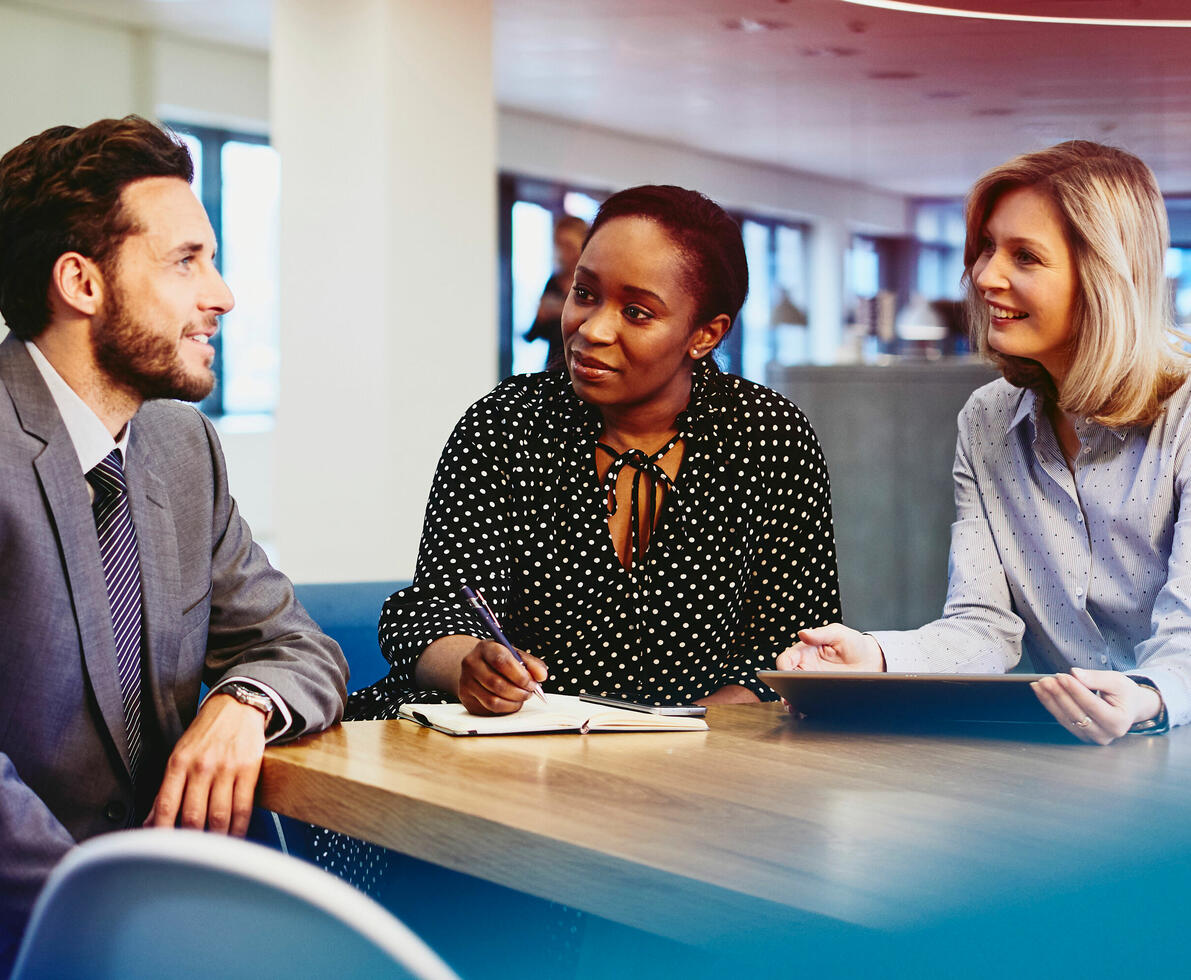 an image of three colleagues chatting while sitting down and one is taking notes