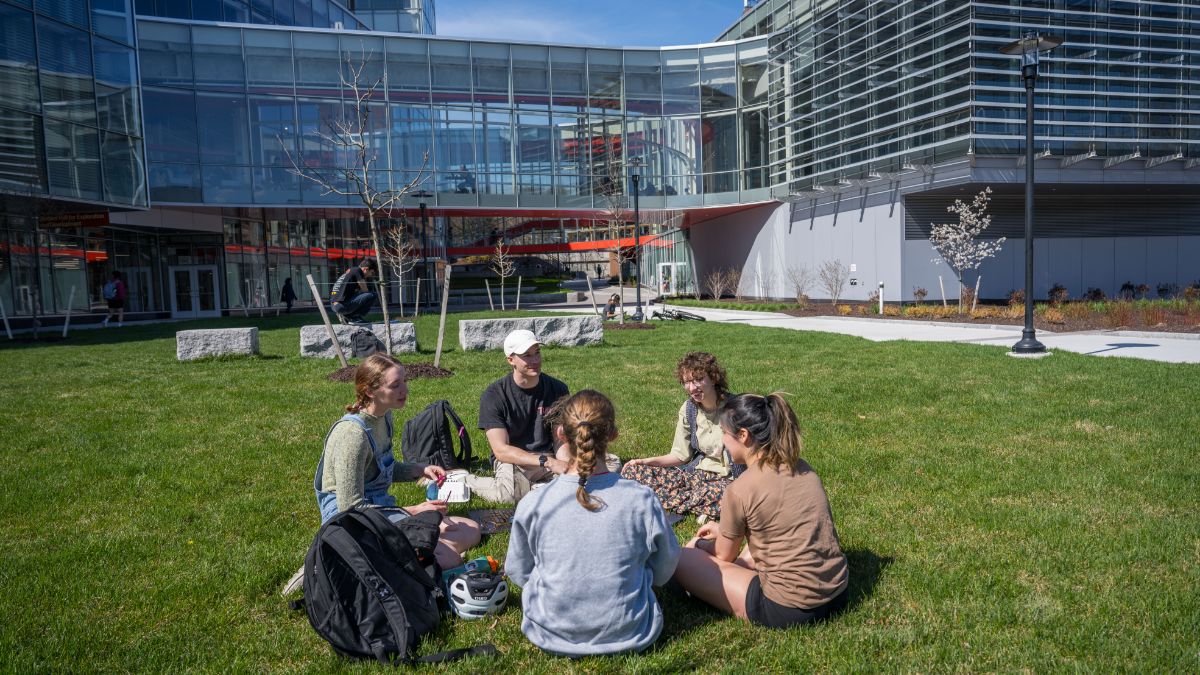 A group of students sitting on a grassy field in front of a modern glass building on a sunny day.