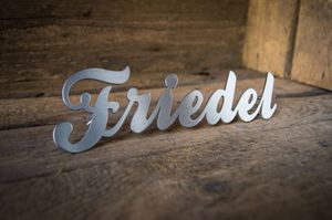 Schriftzug Friedel aus gebürstetem Edelstahl vor Holzwand