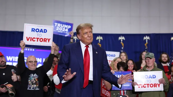 A person in a suit stands in front of a crowd of people holding signs that say "Trump 2024" and "Road to Victory."