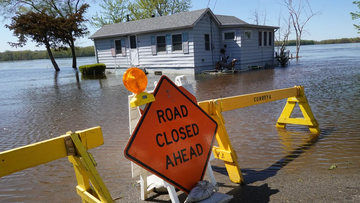 An orange sign reading "Road Closed Ahead" in a flooded road in front of a house.