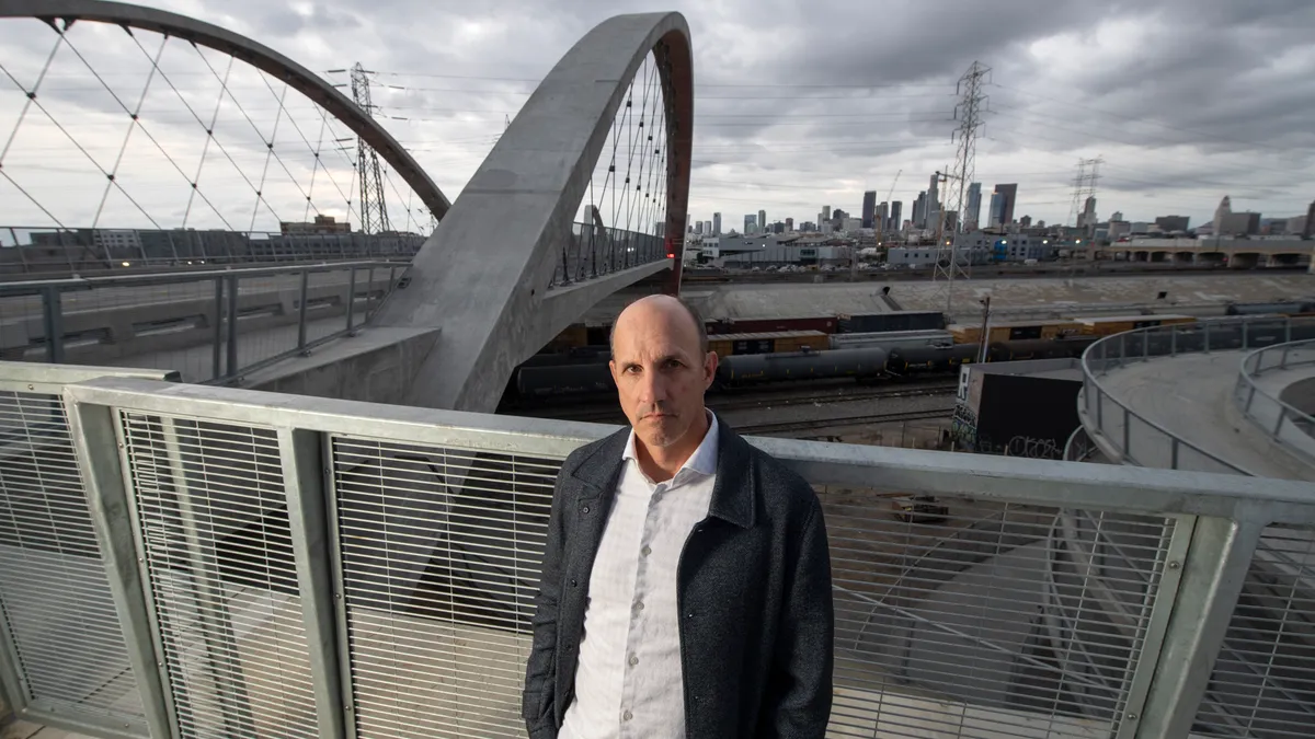 A person in a button down shirt and jacket stands in front of a bridge and city skyline.