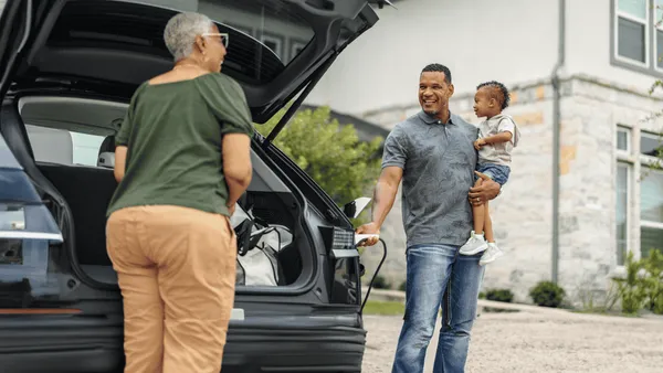 A family packs their car for a trip, with a man holding a child and a woman loading the trunk.