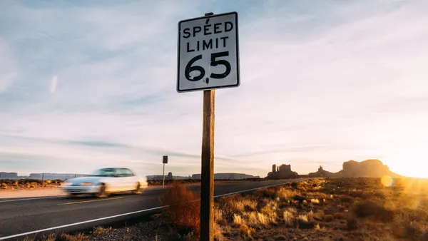 A car passes a sign reading "Speed Limit 65" on a desert highway.