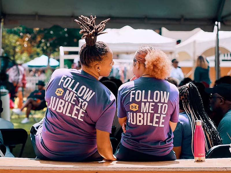 Two women in Jubilee t-shirts sitting facing a stage of performers.