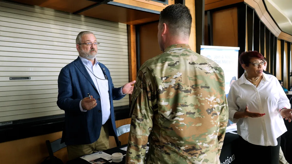 A man in a blue blazer speaking with a active duty service member in a green camouflage uniform.