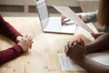 Close up photo of three people in a job interview. One person holds the candidates CV while the others have their arms on the table.