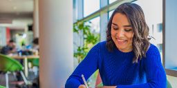 Female student with a bright blue jumper is seen studying a desk in a well-lit campus space, she is drawing on a tablet.